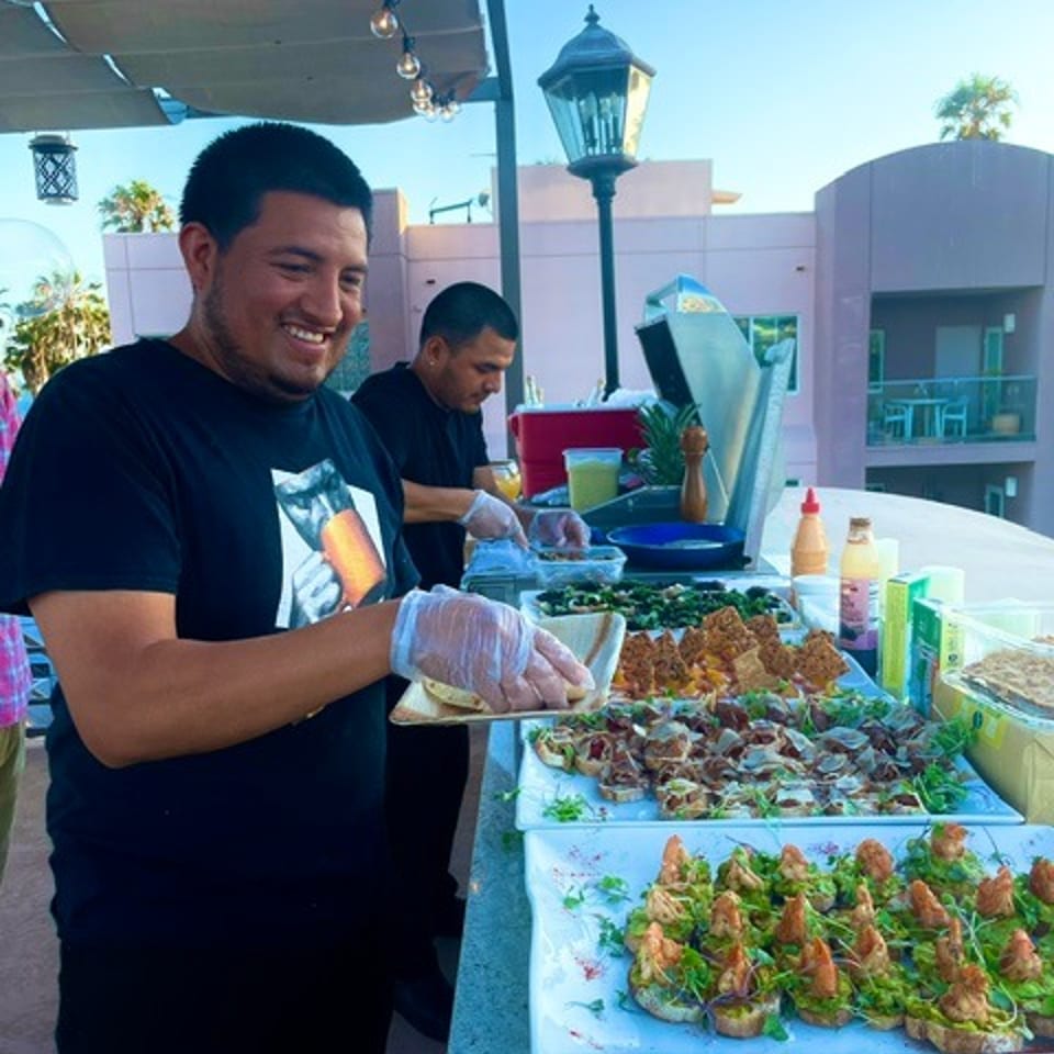 Two men prepare colorful seafood dishes at an outdoor food stall, with trays of garnished food and condiments on the counter.
