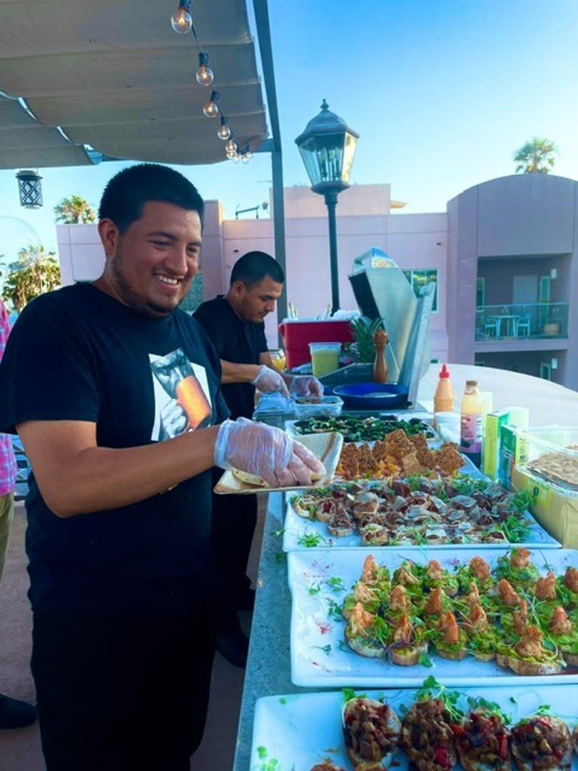 Smiling server with gloves presents appetizers from a vibrant outdoor buffet spread.