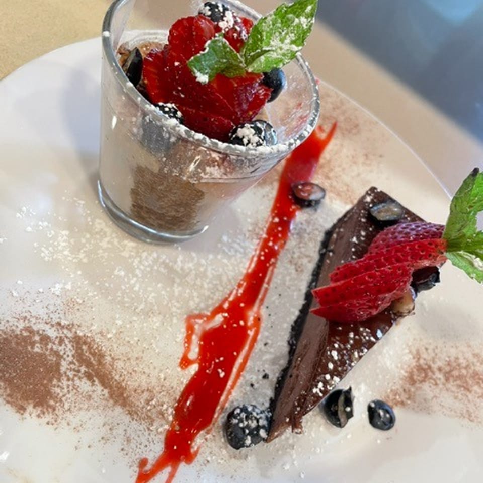 Chocolate dessert with berry-topped mousse cup, chocolate slice, mint leaves, powdered sugar, and raspberry sauce on a white plate.