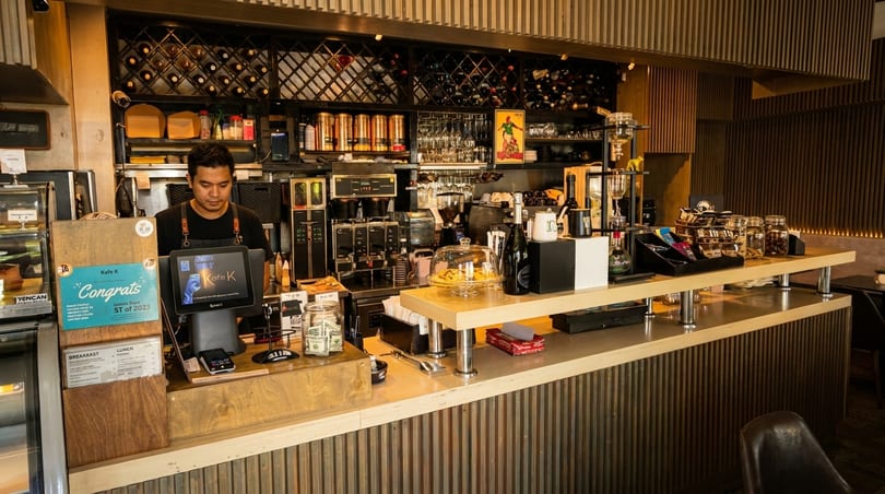 A barista stands behind a well-stocked coffee bar with wine racks and espresso machines.