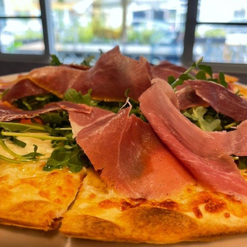 Thin-crust pizza topped with cured ham, leafy greens, and olive oil, served near a bright restaurant window.