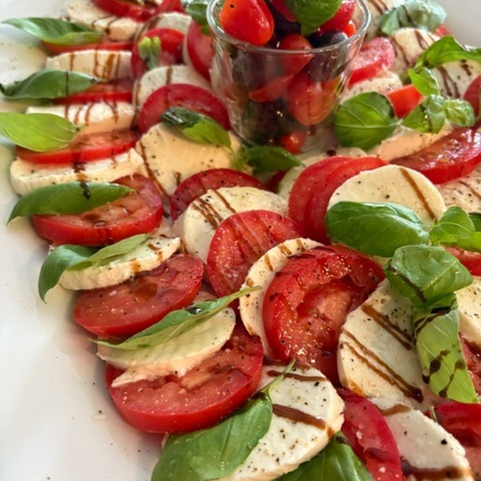 Caprese salad with sliced tomatoes, mozzarella, fresh basil, balsamic drizzle, and a glass of cherry tomatoes.