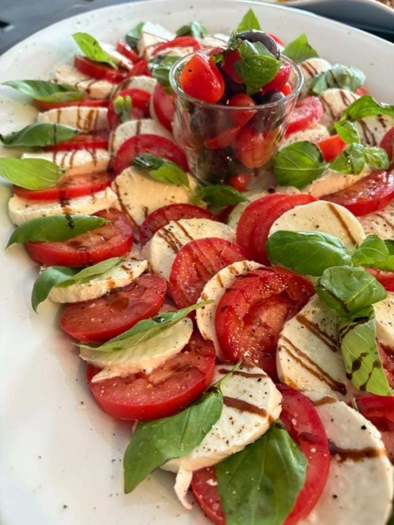 Caprese salad with sliced tomatoes, mozzarella, basil, balsamic glaze, and a central glass of cherry tomatoes.