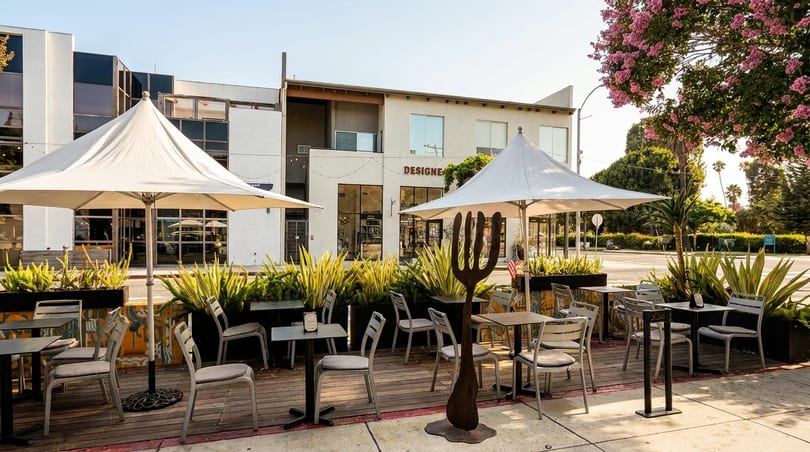 Outdoor patio dining area with tables, chairs, umbrellas, and planters, next to a building.