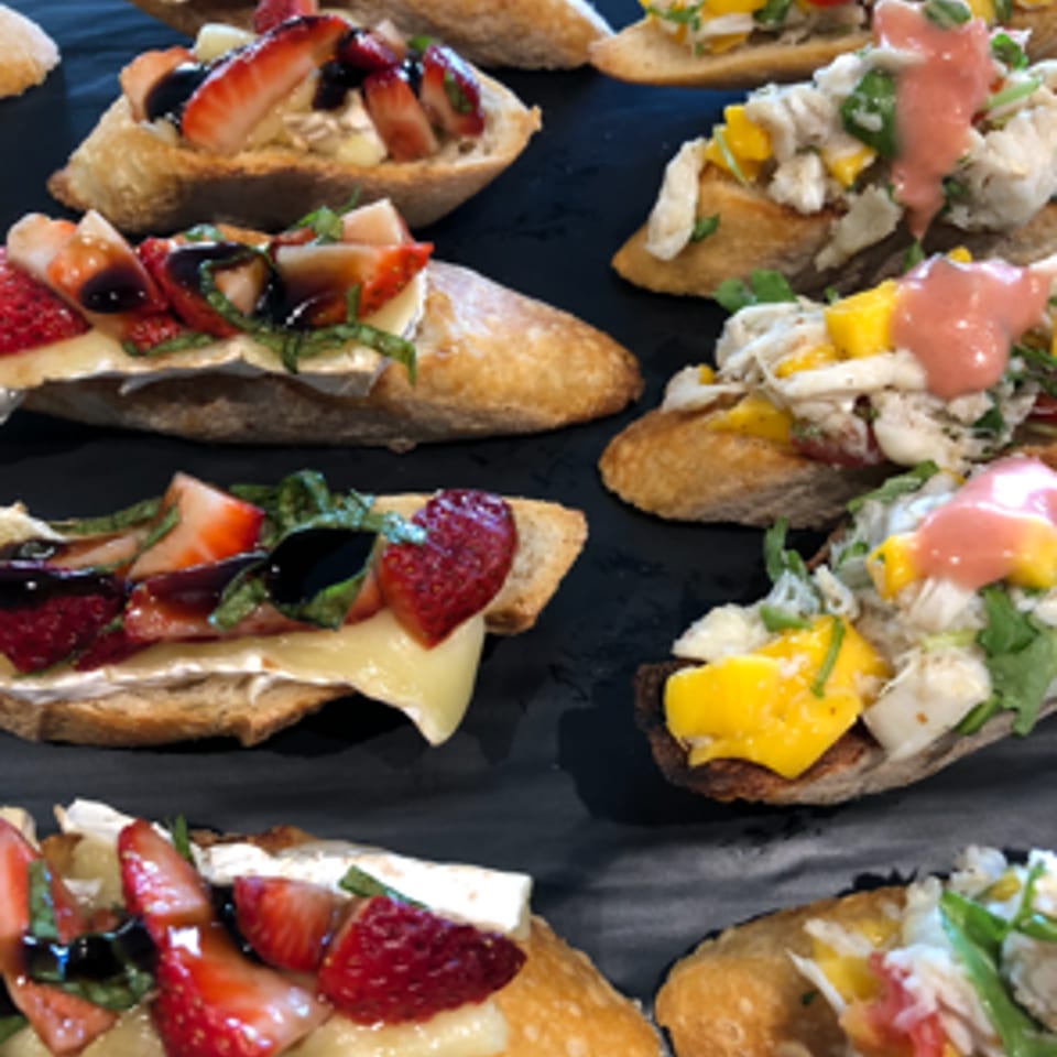 Assorted toasted crostini topped with strawberries, vegetables, creamy cheese, herbs, and pink sauce on a dark platter.